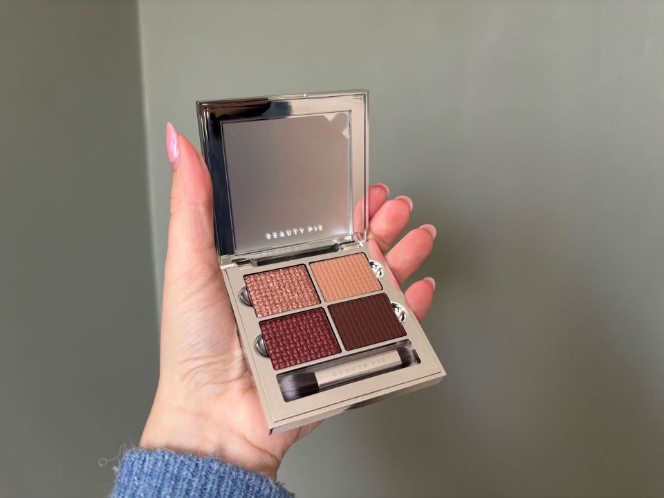 Hand holding open Beauty Pie eyeshadow palette with four shades and a small brush.