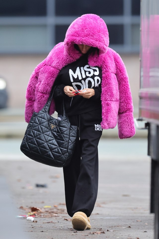 Woman walking while wearing a pink fuzzy hooded jacket, black Miss Dior sweatpants, and carrying a black quilted bag.