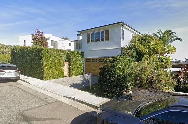 Two-story white house with a wooden garage door, partially obscured by hedges.