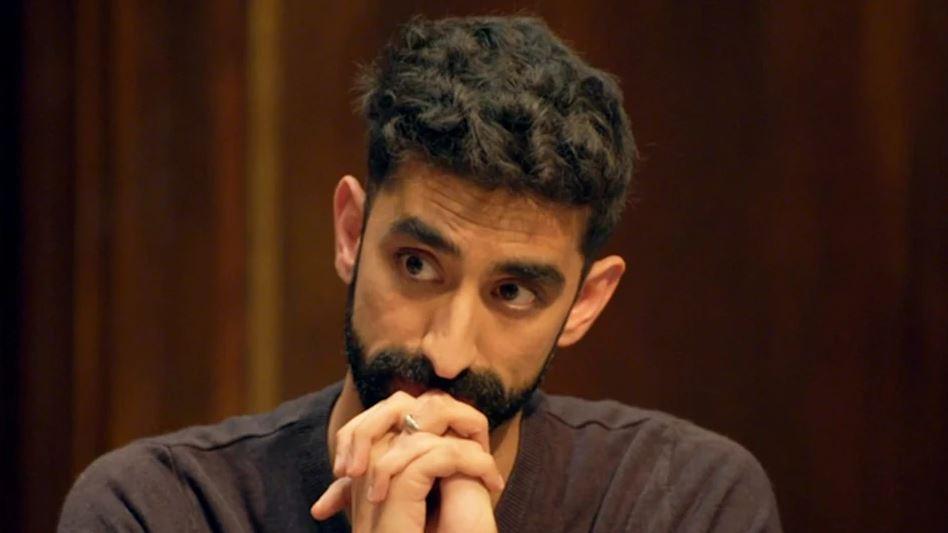 A man with dark curly hair and a beard sits looking pensive.