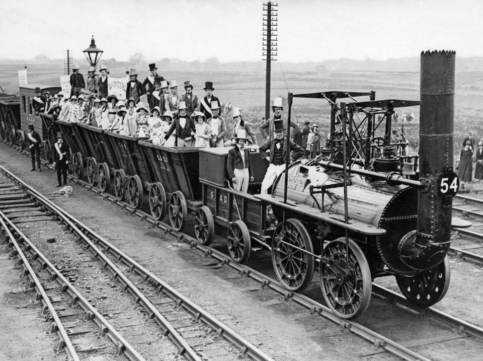 A steam train carrying many passengers in top hats and dresses.