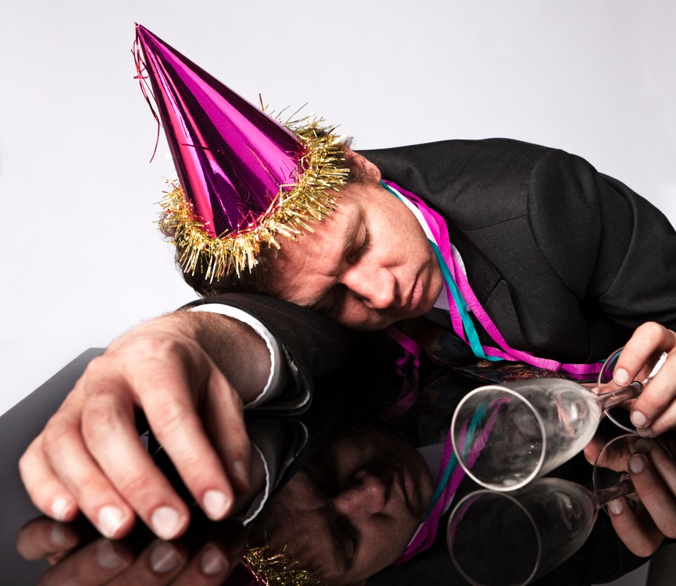 A businessman asleep at a party, wearing a party hat and holding an empty champagne flute.