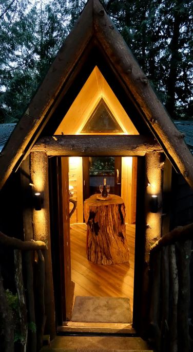 Lit interior of an A-frame treehouse with a wooden stump table and wine.