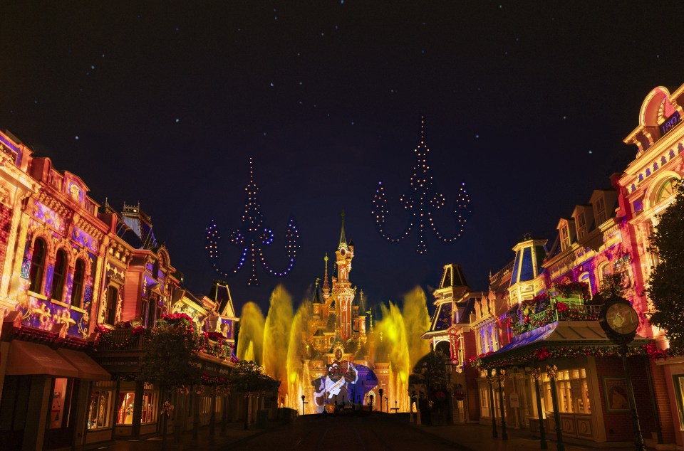 Nighttime view of Disneyland Paris with light projections on buildings and a drone light show above.