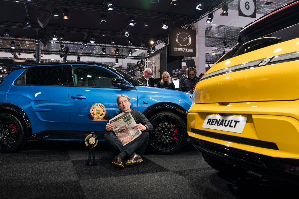 Man sitting between a blue and yellow car, holding a newspaper.