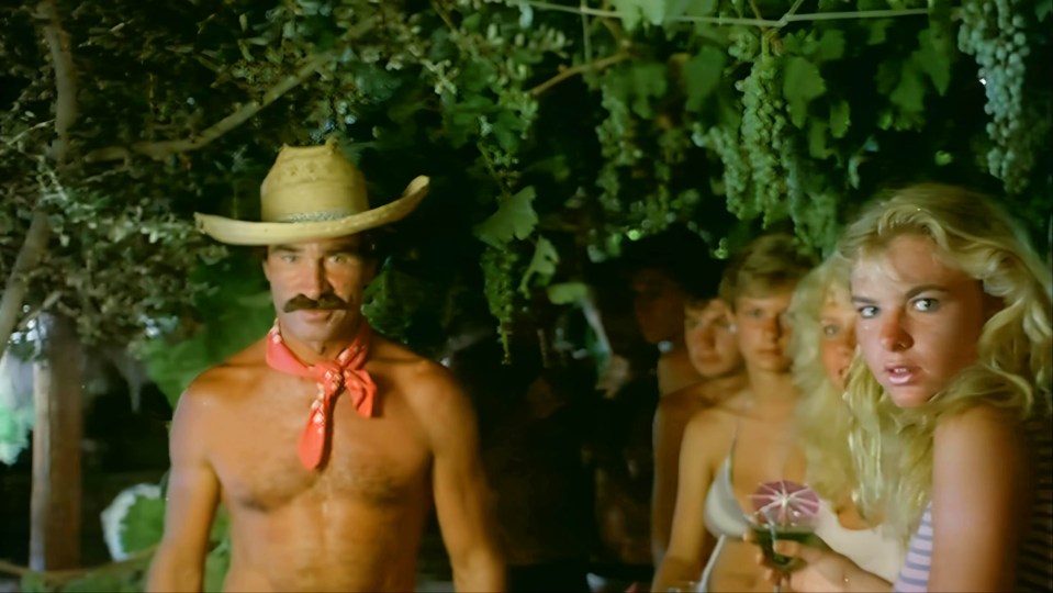 A shirtless man in a cowboy hat and red bandana stands in front of a group of young women under a grapevine.