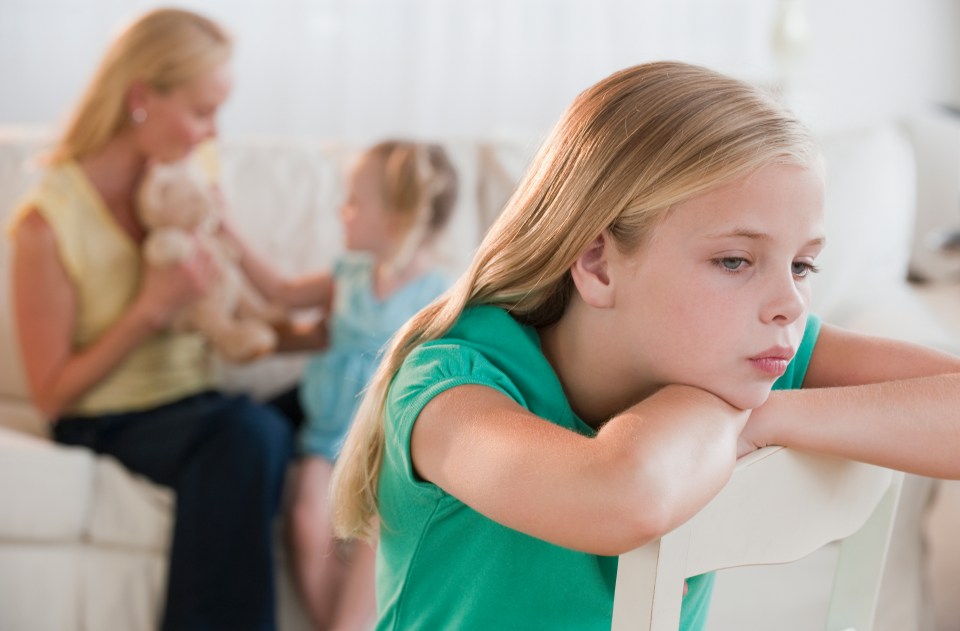 Sad girl sitting in a chair, her mother and younger sister in the background.