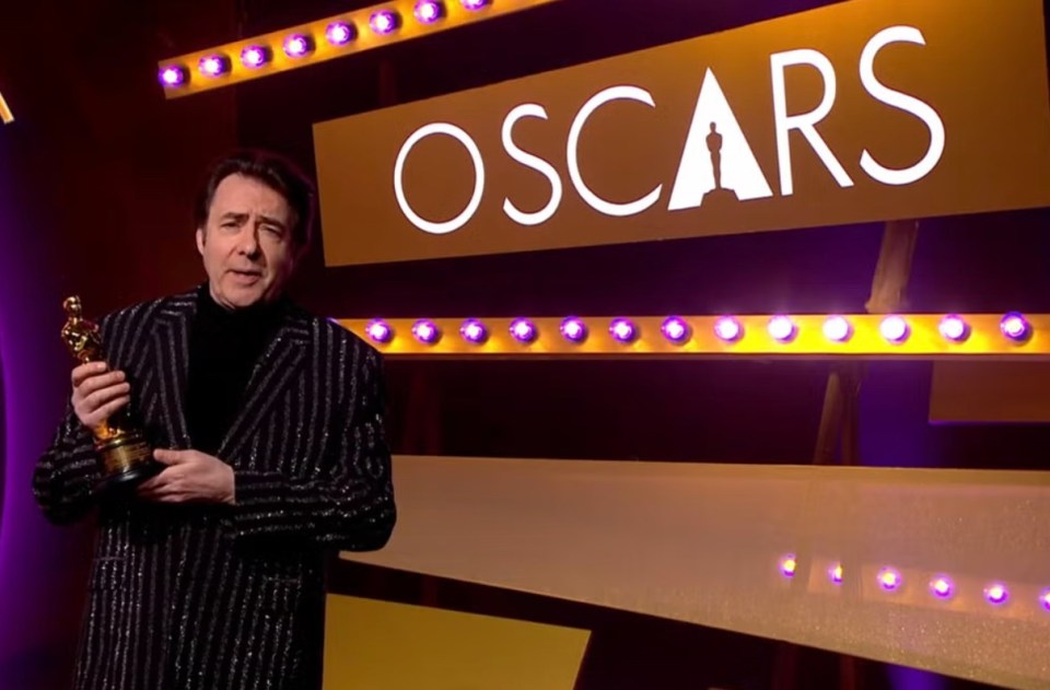 Jonathan Ross holding an Oscar statuette in front of an Oscars backdrop.