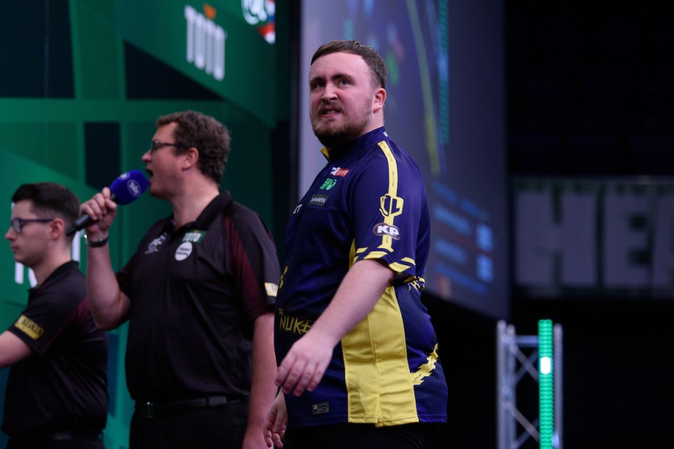 A darts player in a dark blue and yellow shirt stands on stage.