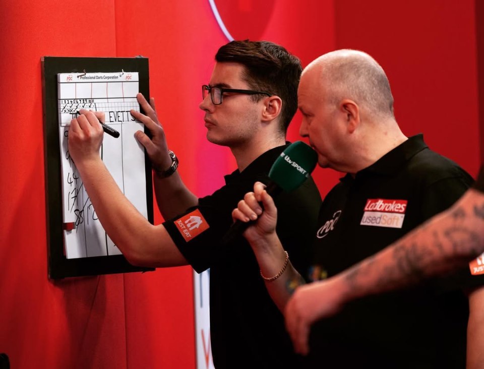 A darts scoreboard being updated by a player, with a commentator holding a microphone nearby.