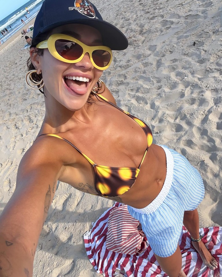 Woman on beach in bikini top and shorts taking a selfie.