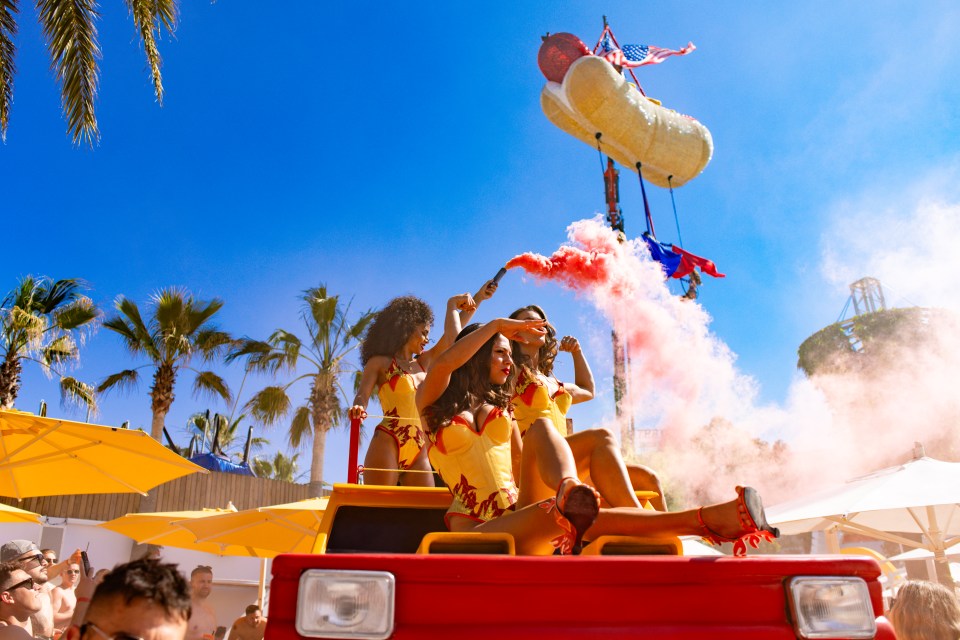 Three women in yellow and orange costumes on a red vehicle release pink smoke.