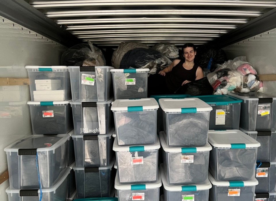 Woman sitting amidst numerous plastic storage bins full of clothing in the back of a truck.