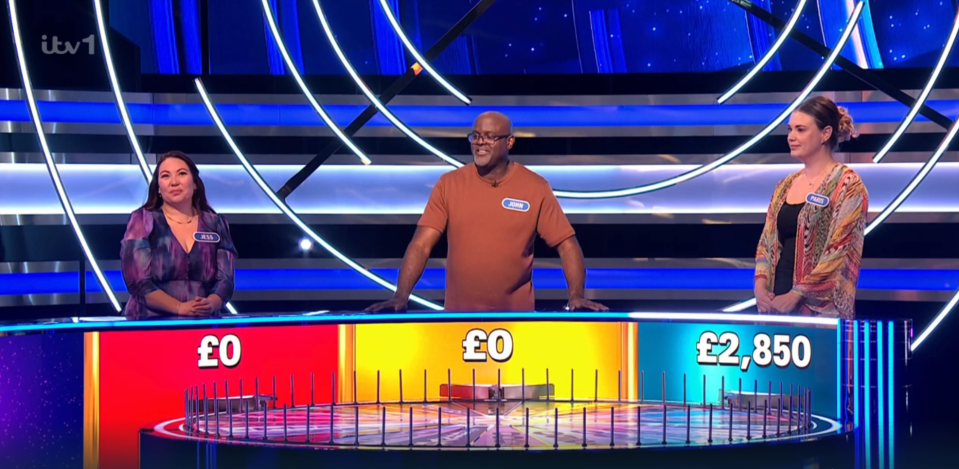 Three contestants stand at a game show podium with a spinning wheel showing £0, £0, and £2,850.