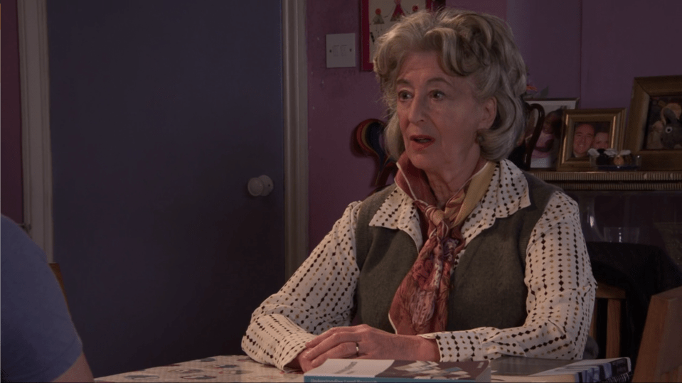 Older woman in a patterned shirt and vest sits at a table, speaking.