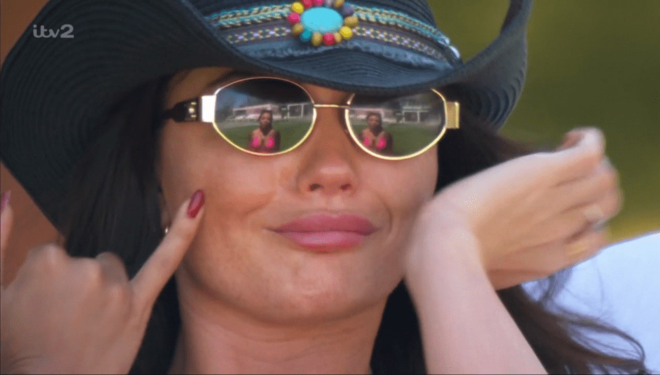 Woman wearing a large hat and sunglasses; reflection in sunglasses shows two women in bikinis.