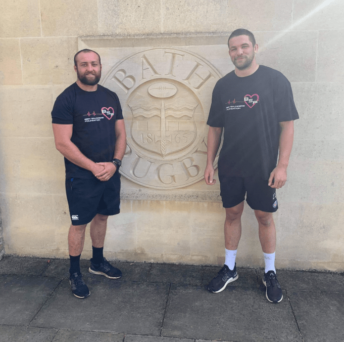 Two men in black Bath Rugby t-shirts stand in front of a Bath Rugby emblem.