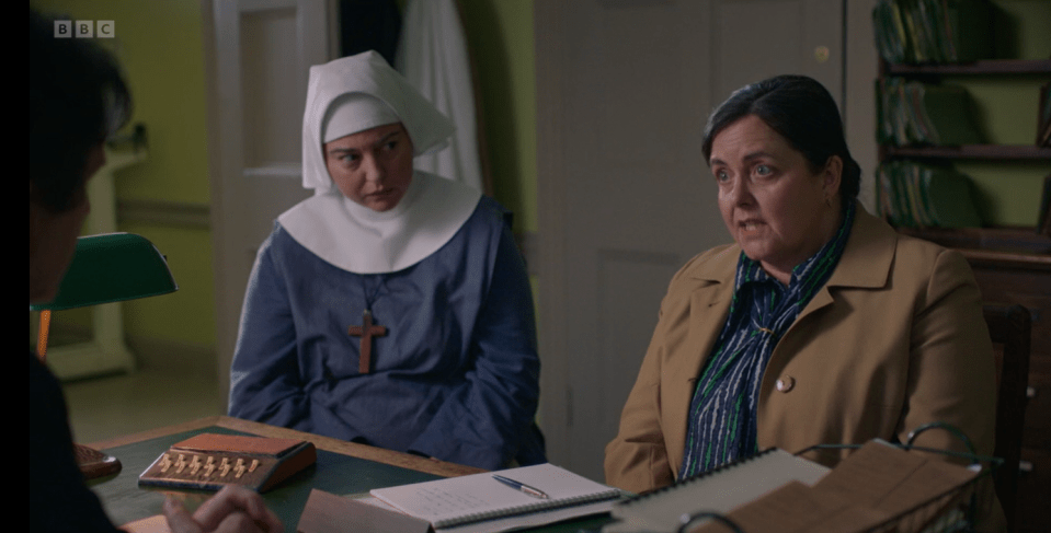 Screenshot of a scene from a television show, showing a nun and a woman in a tan coat sitting at a table talking to a person off-camera.