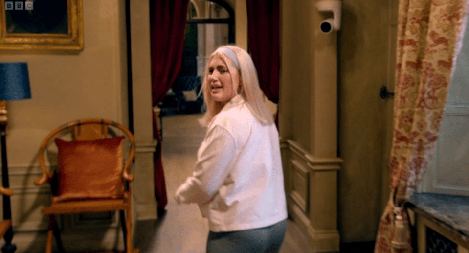 Screenshot of a blonde woman walking away from the camera in a hallway.