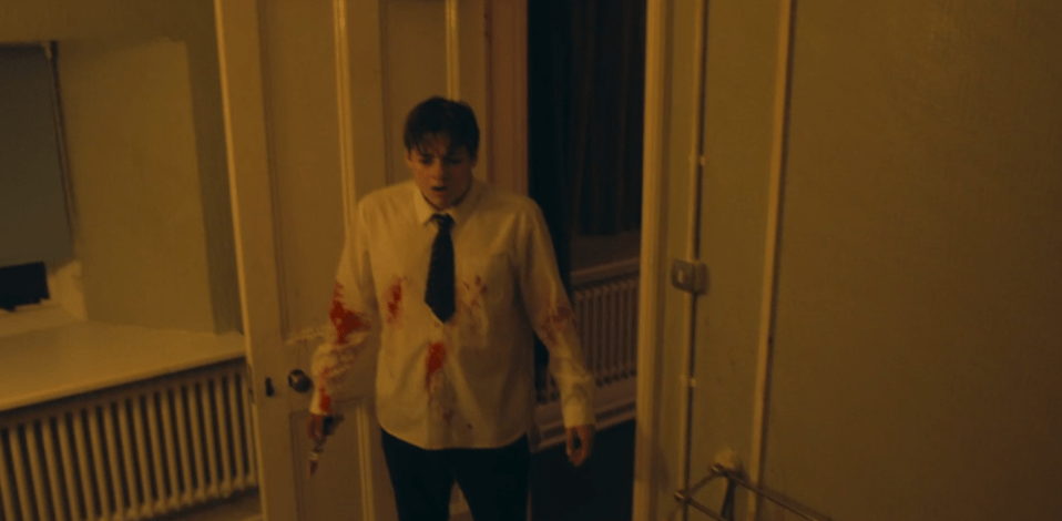 A blood-splattered young man in a white shirt and tie stands in a doorway.