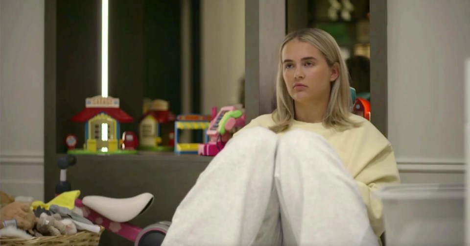 Woman sitting with legs crossed, looking pensive; toys visible in the background.