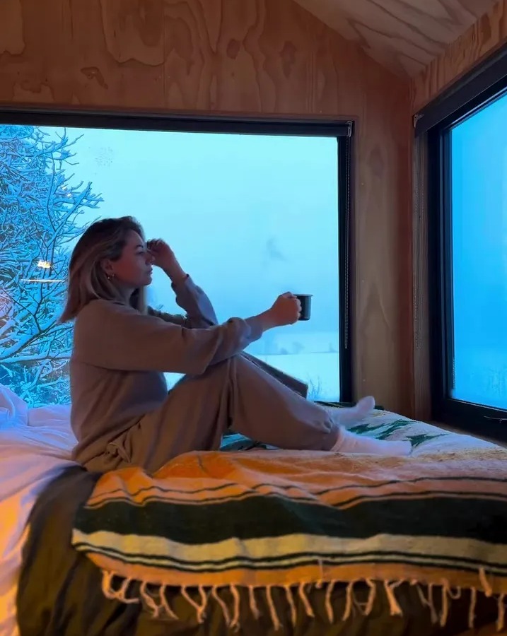 Woman sitting on a bed in a cabin, enjoying a hot drink while looking out at a snowy landscape.