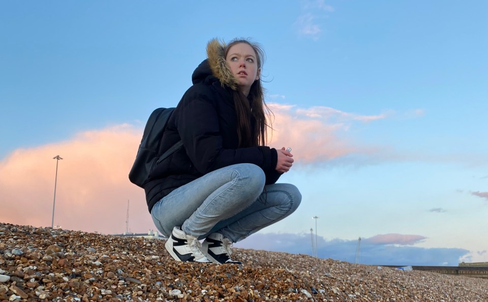 Woman crouching on a pebble beach, looking up at the sky.