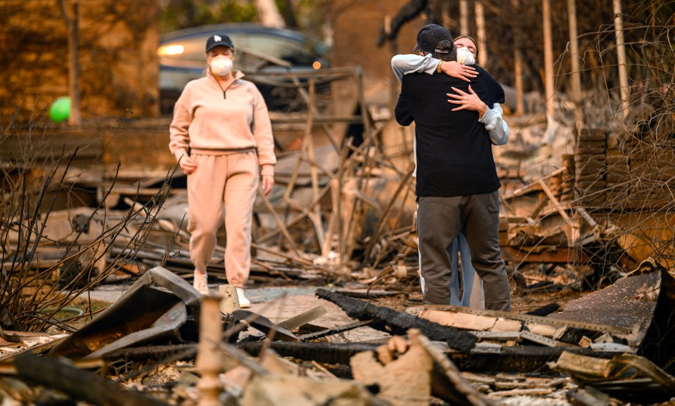 A man and woman embrace amidst the debris of a fire-damaged home.  A woman walks in the background.