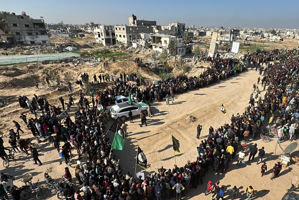 Aerial view of a large crowd gathered in a war-torn area, with flags and a vehicle present.