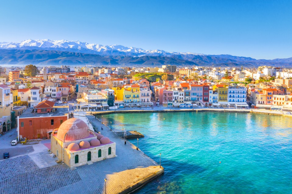 Aerial view of Chania, Crete, showing the harbor, lighthouse, and snow-capped mountains.