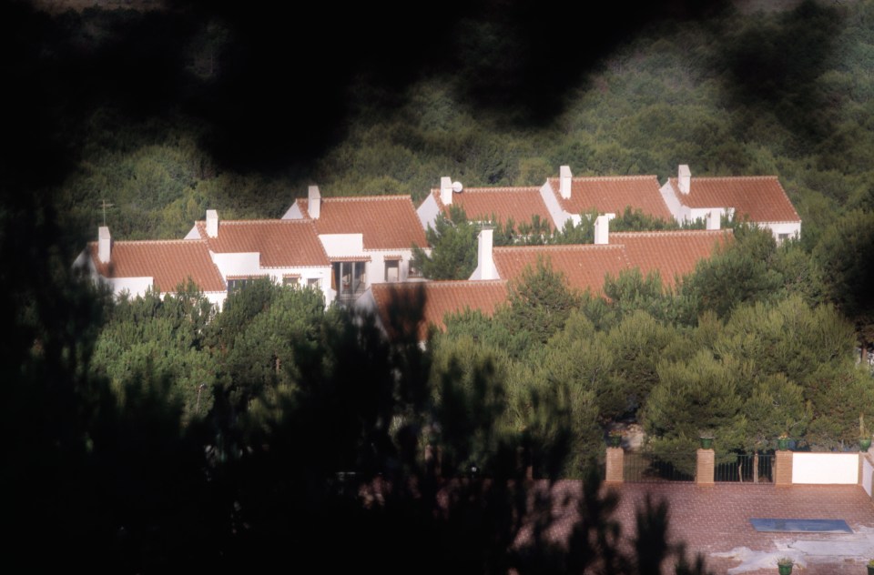 Set of white buildings with red tile roofs, partially obscured by trees.