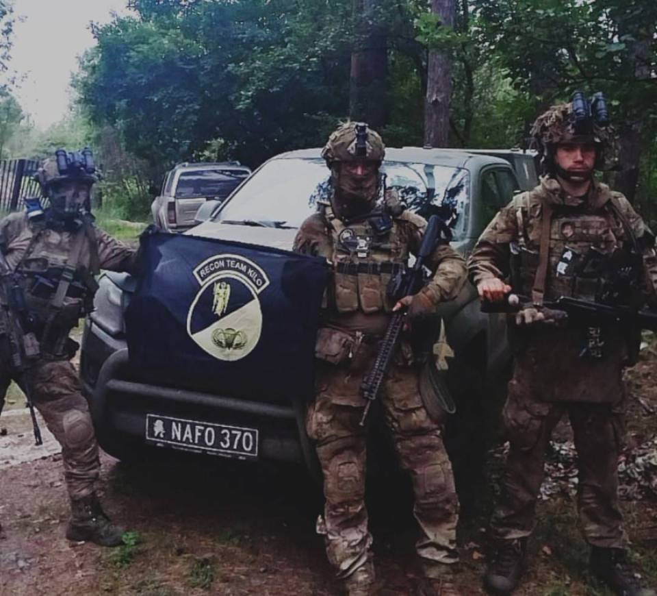 Photo of a group of soldiers in camouflage uniforms posing with a flag in a wooded area.