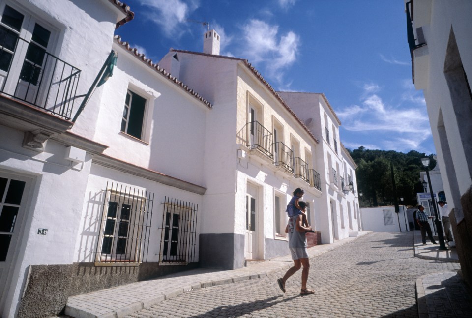 A man carrying a child on his shoulders walks down a cobblestone street lined with white buildings.