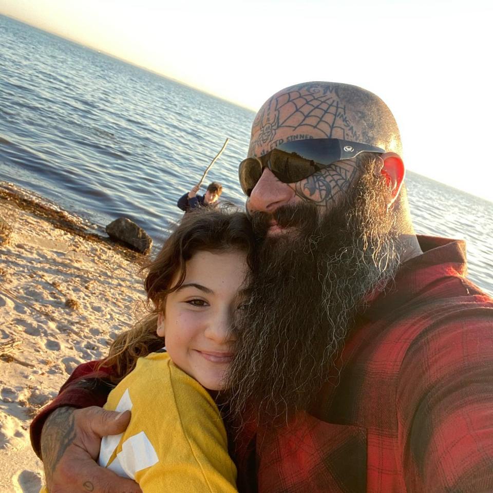 A man with extensive tattoos hugs a young girl on a beach.