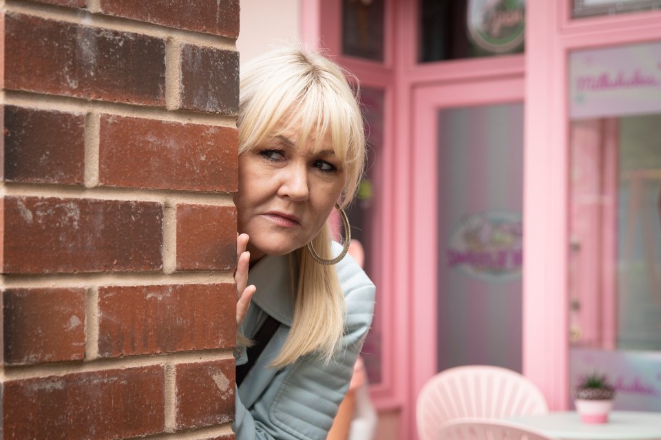 Beth Tinker from Coronation Street spies from behind a brick wall.