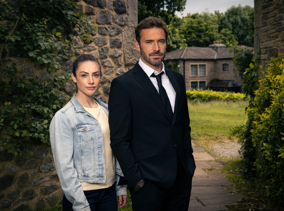 A woman in a denim jacket and a man in a suit stand outside a stone building.