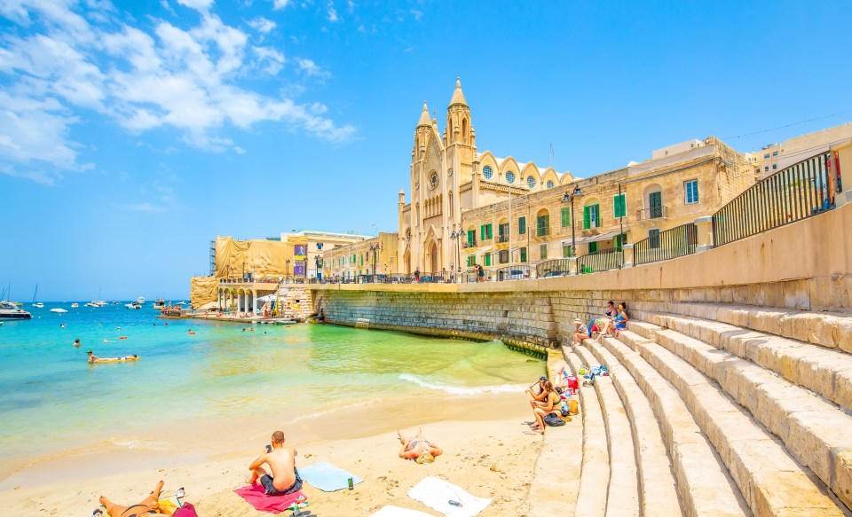 Balluta Bay beach in St. Julian's, Malta, with people sunbathing and swimming near a church.