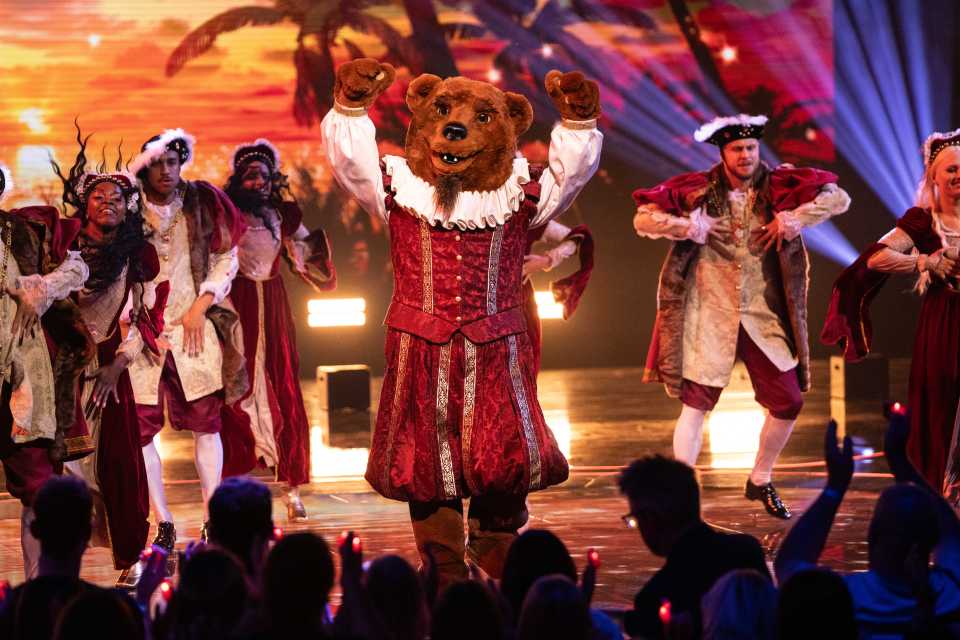 Bear in a red costume performing on stage with backup dancers.