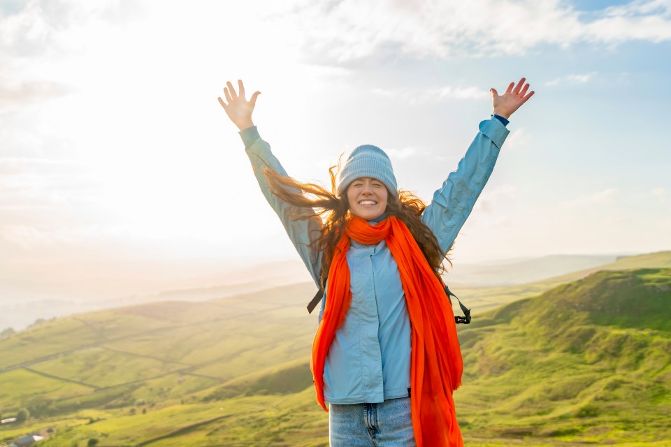 A joyful young woman with arms raised stands on a hilltop.