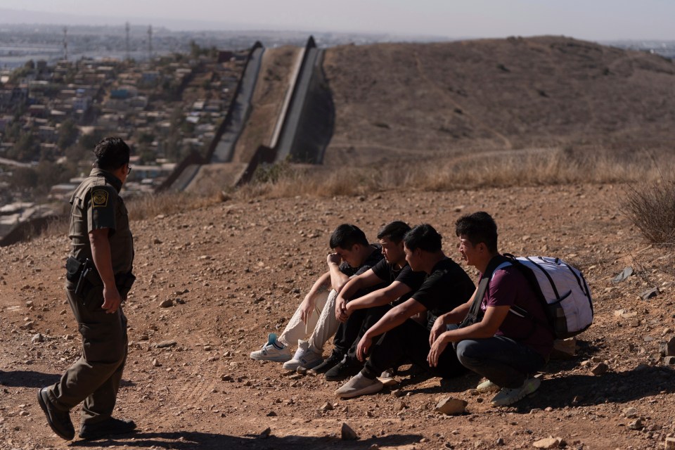 Border patrol agent and four detained migrants near a border wall.