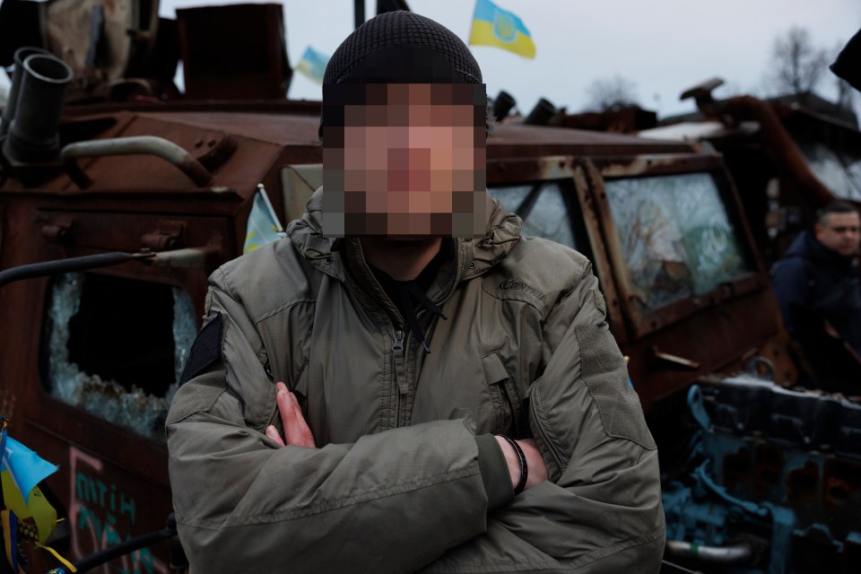 British frontline medic with face obscured, standing in front of damaged military vehicle.