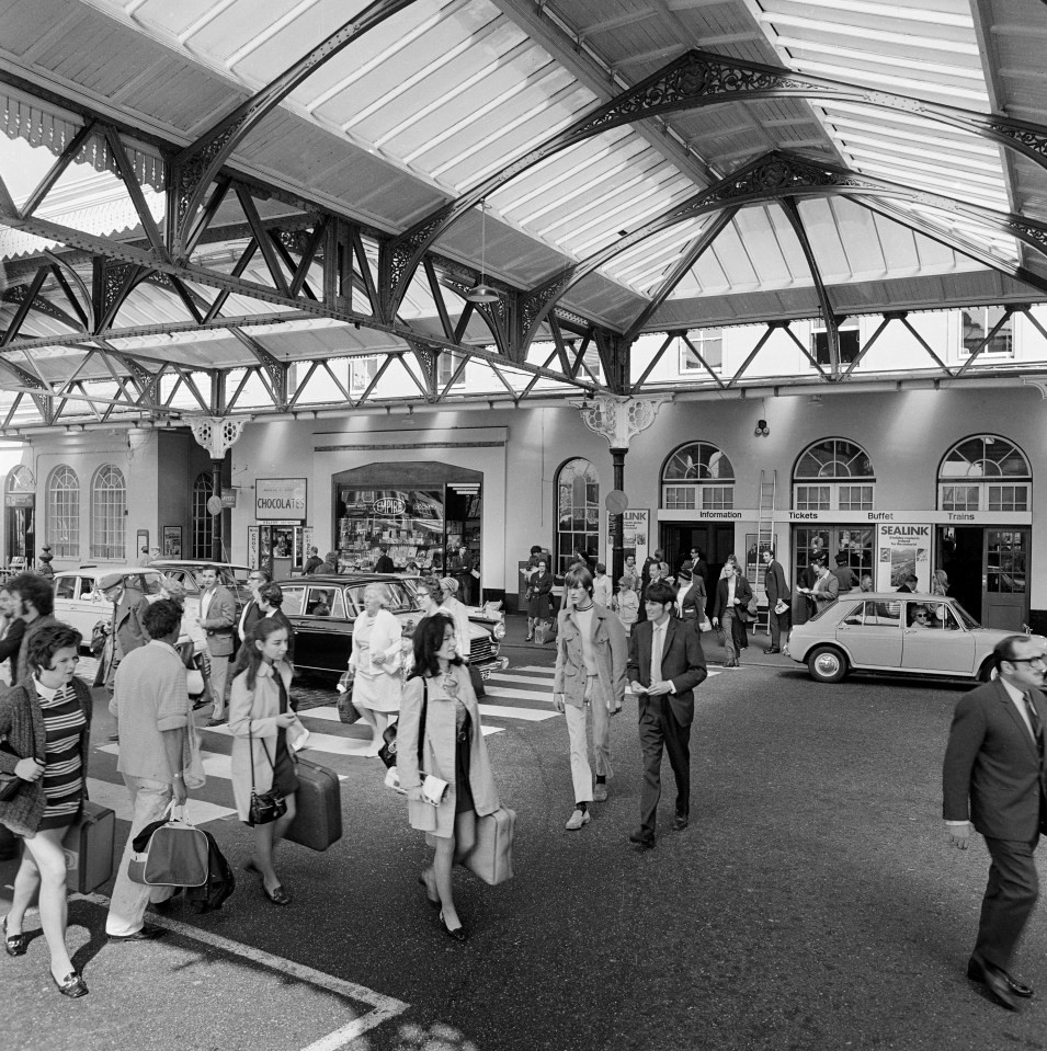 Black and white photo of people with suitcases crossing a street in front of a train station in the 1960s.