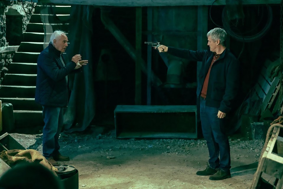 A man points a gun at another man in a dark barn.