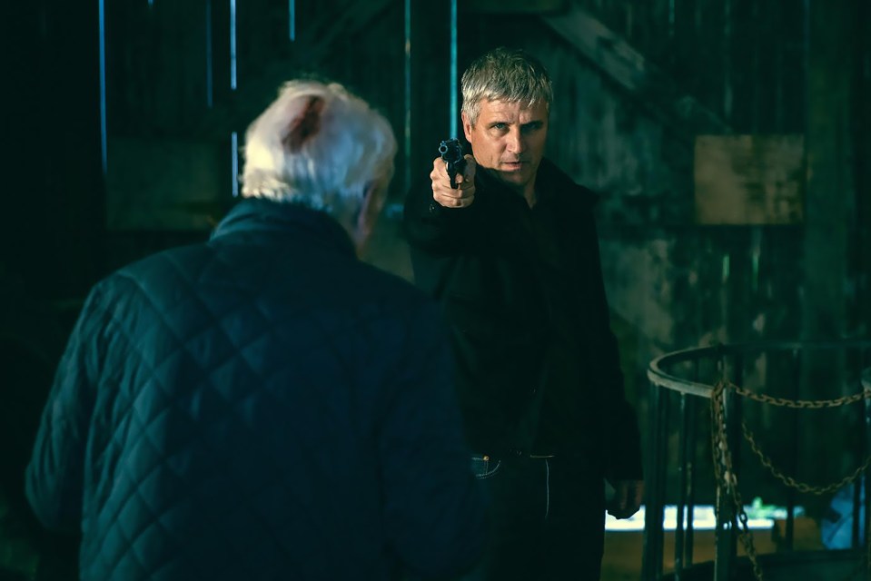 A man points a gun at another man in a barn.