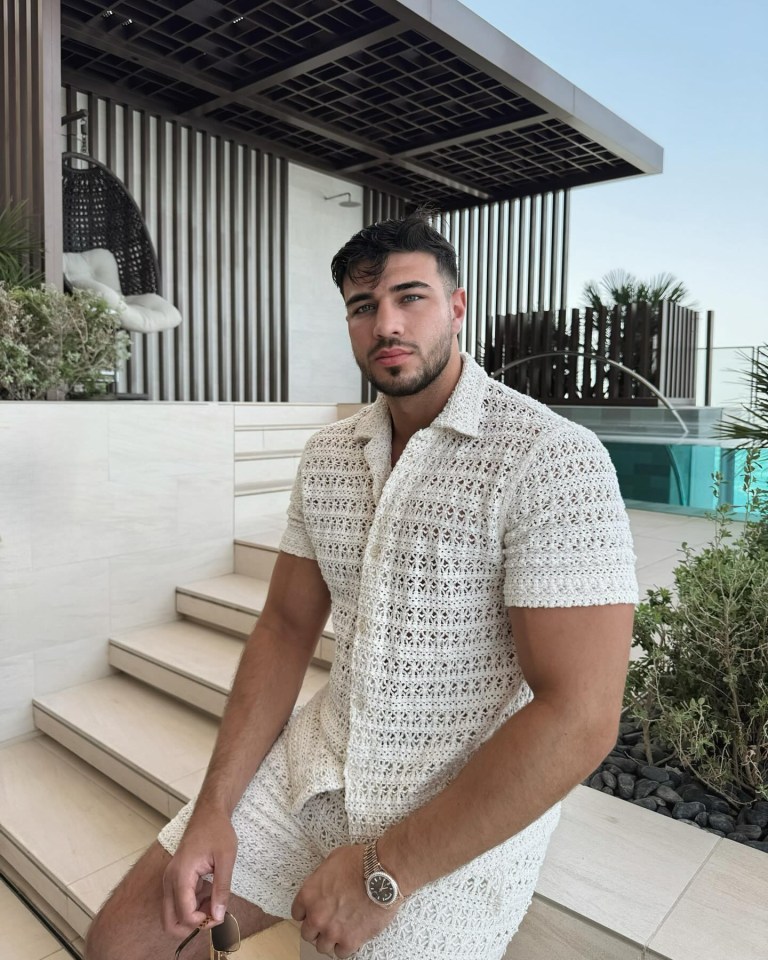 Man in crocheted cream-colored shirt and shorts sitting on steps by a pool.