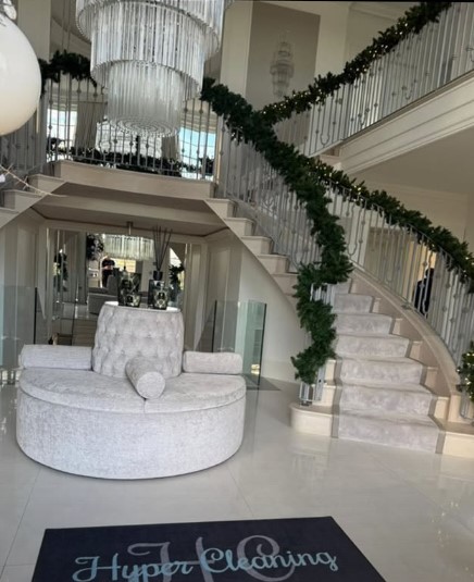 Interior of a large home with a grand staircase decorated with garland, a large chandelier, and a circular light grey sofa.