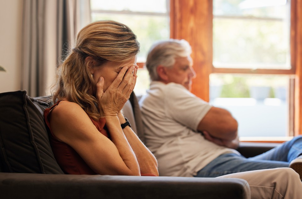 A senior woman cries while sitting on a couch next to her husband, who is sitting with his arms crossed.