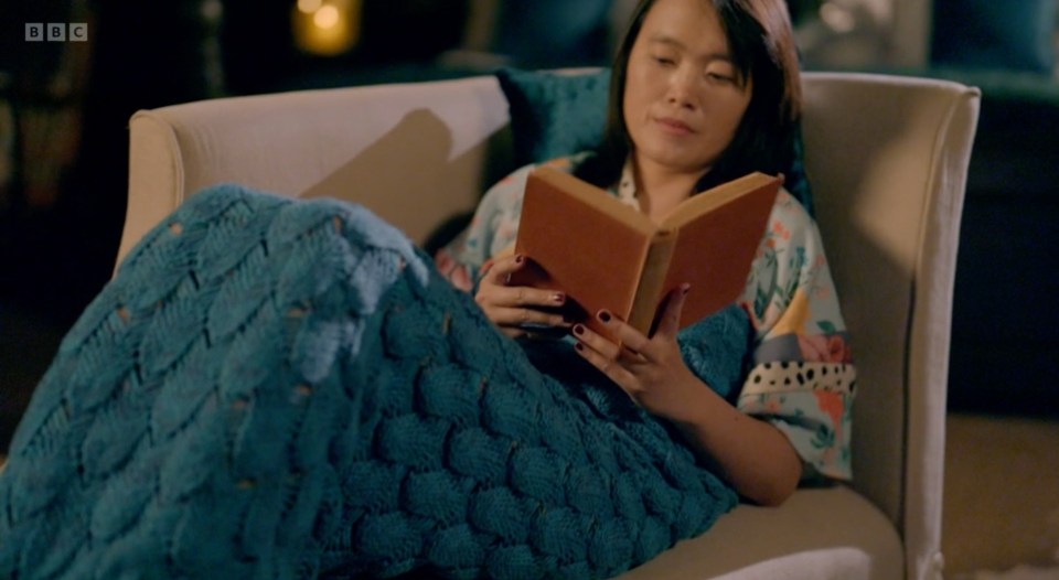 Woman relaxing on a couch, wrapped in a teal blanket, reading a book.