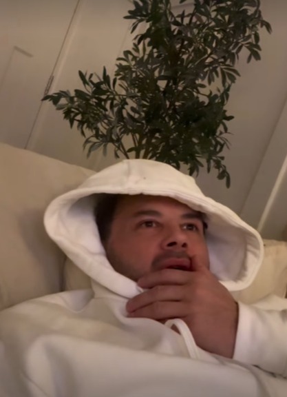 Ryan Thomas in a white hoodie, a plant on his head, looking pensive.