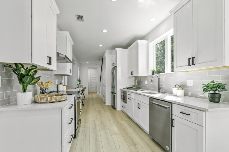 Interior view of a narrow, modern kitchen with white cabinets and gray tile backsplash.